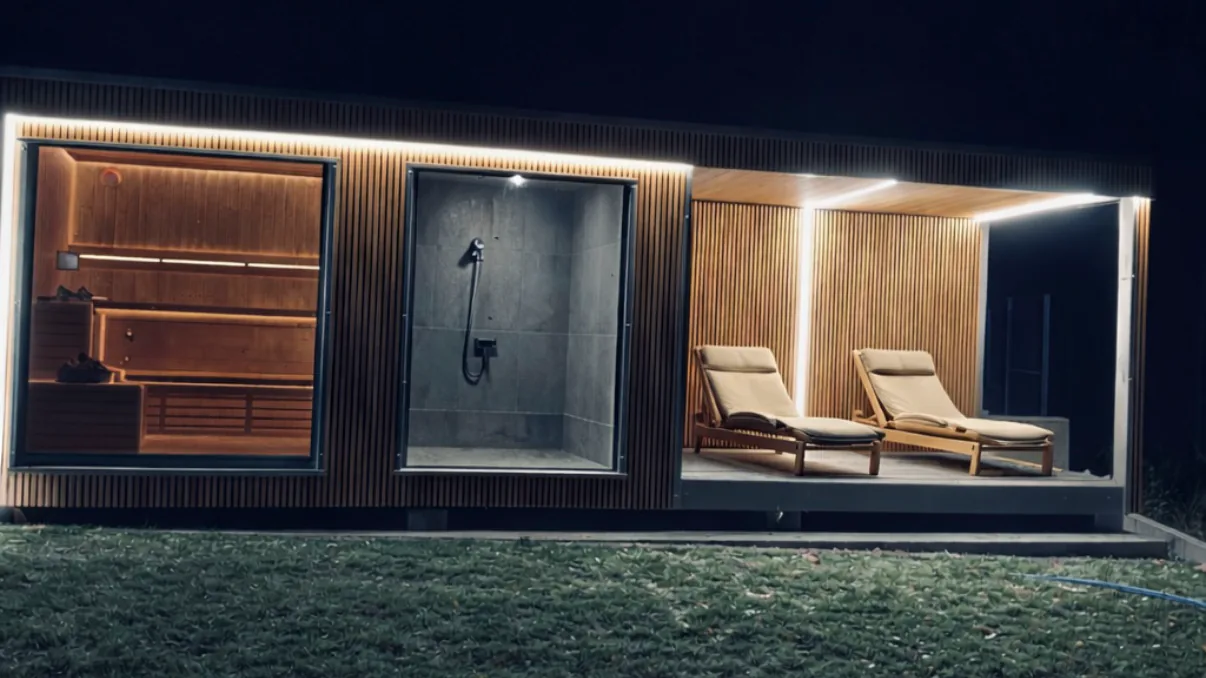 Sauna ogrodowa nocą: oświetlona kabina, prysznic i zadaszona strefa relaksu z leżakami